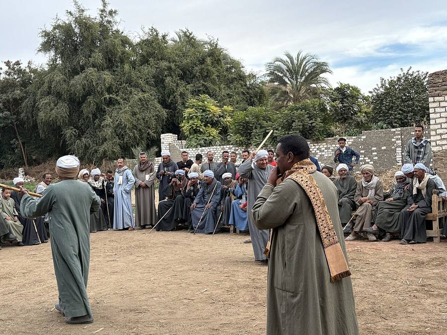 Two men in long robes face each other holding long sticks in a tahtib stick-fighting display at a festival in Luxor, watched by a crowd